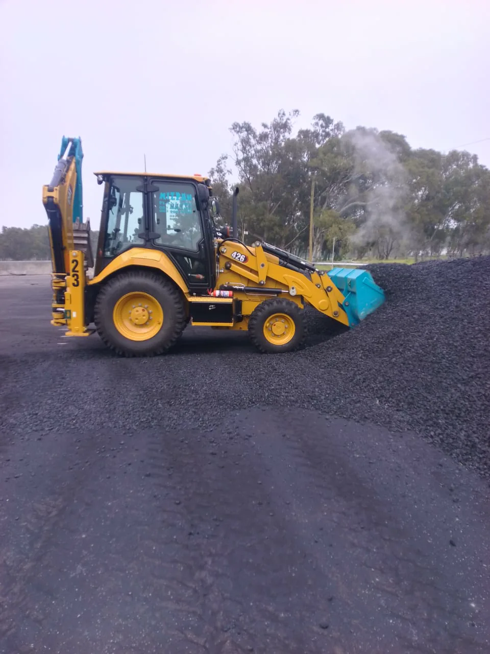 Mizpah Plant Hire TLB loading dark aggregate at a material yard